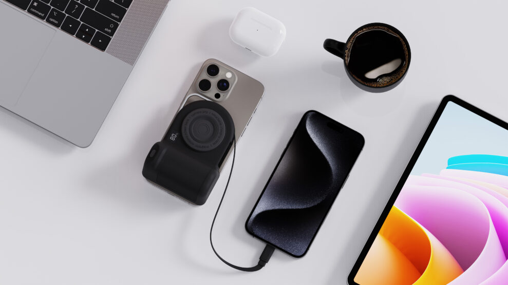 Flatlay av Belkin PowerGrip festet til en sølvfarget iPhone med fire kameralinser, ved siden av en MacBook, et par hvite øretelefoner i ladeetui, en kaffekopp og et nettbrett som viser en fargerik skjerm. Alt ligger på en hvit overflate.